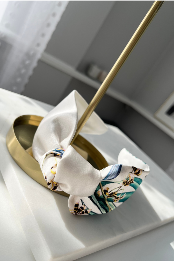 close up image of a white silk headband adorned with a cluster of delicate flowers and lush green leaves on one side, adding a touch of elegance and nature-inspired beauty to the accessory.